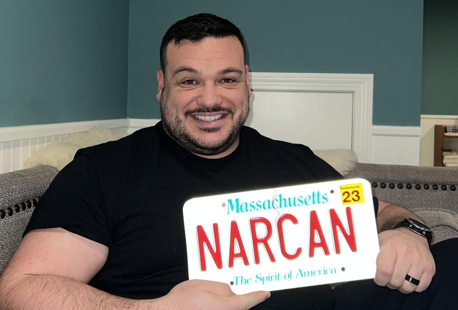 A person sits holding a license plate that says "NARCAN"